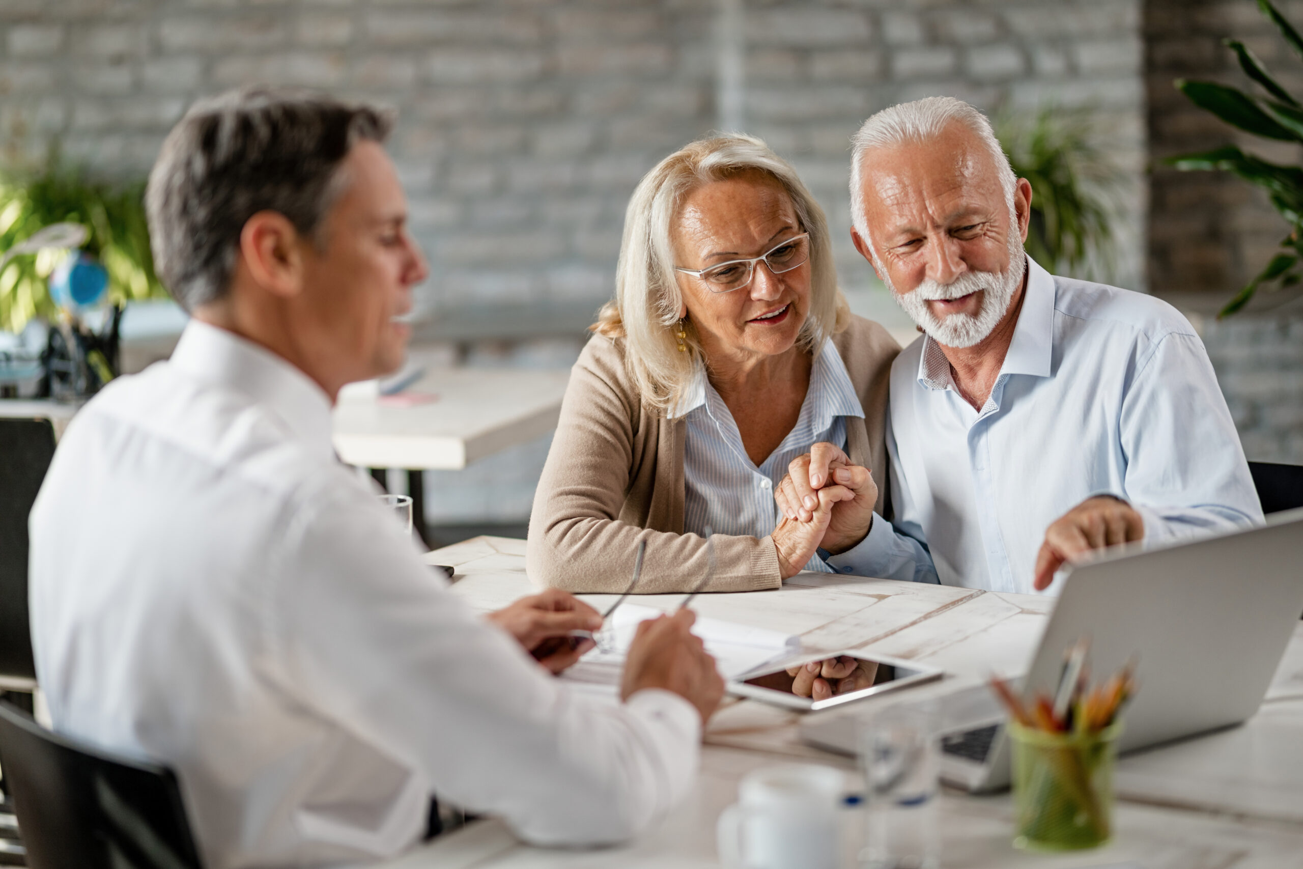 Happy senior couple holding hands and using laptop while having a meeting with financial advisor in the office. Senior man is pointing at something on laptop.